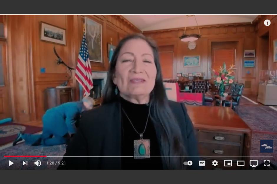 Un empleado pasa arrastr&aacute;ndose, mientras la secretaria del Interior de EE.UU, Deb Haaland, da una entrevista en vivo. (Foto: captura de pantalla)